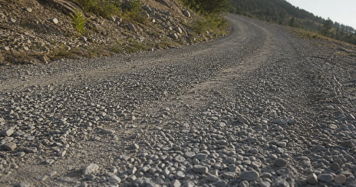 Wide gravel service road with angular gray stones, compacted wheel bands, loose shoulders, and believable low-angle surface relief